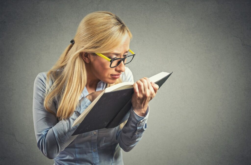 A woman with yellow and black framed glasses squinting and looking at a book in close range.