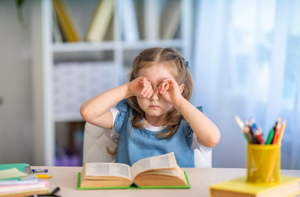 A child rubs their exhausted eyes while trying to read a book.