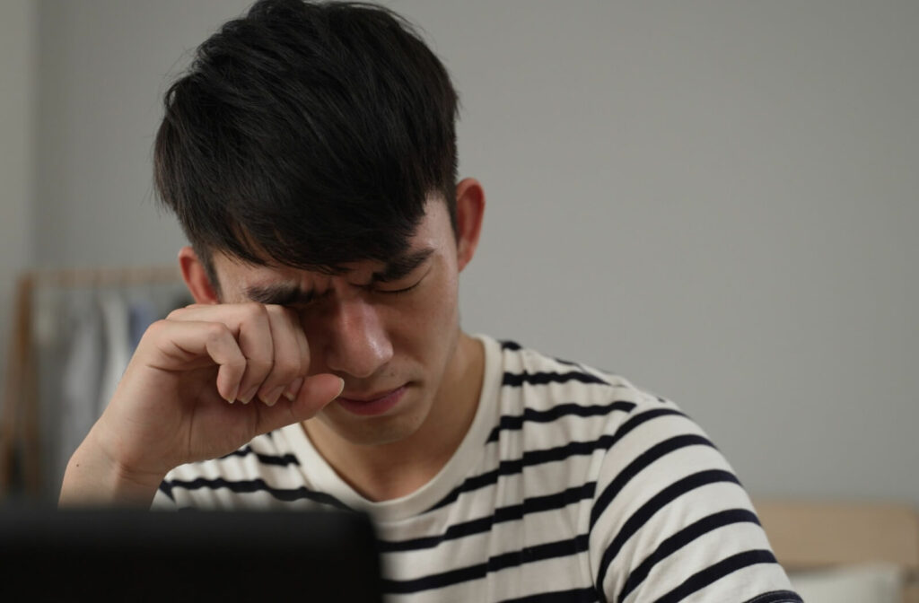 A young man rubs at his dry eyes while sitting in front of his laptop.