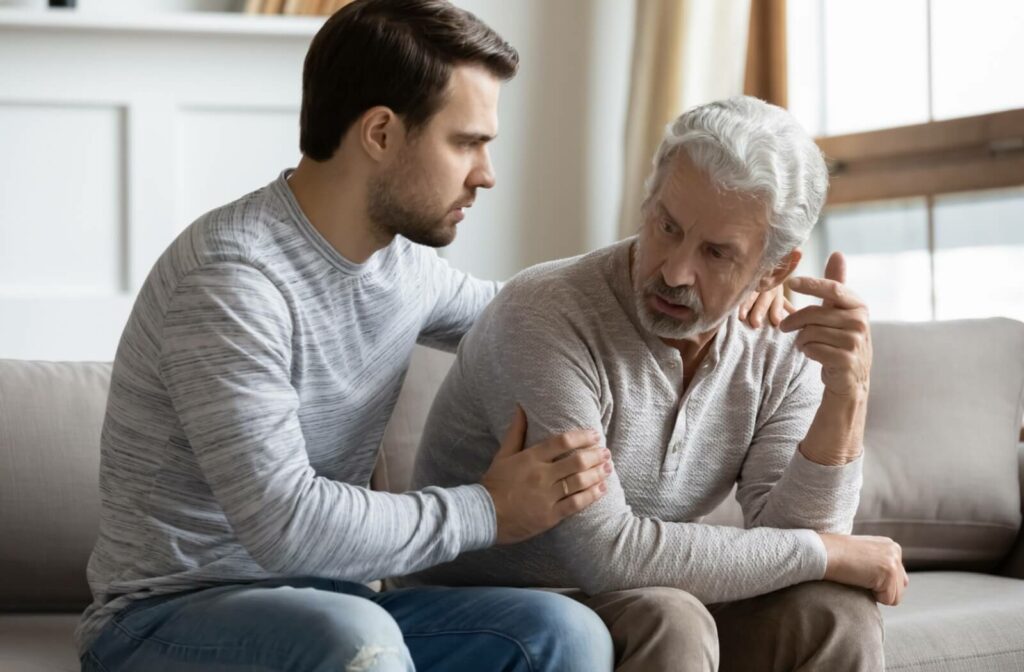 A concerned younger man gently supports an older man sitting on a couch as they discuss an important family health matter