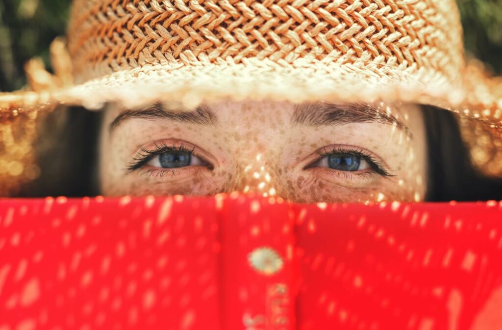 A freckled individual’s blue eyes are framed by a straw, overbrimmed hat and a red book covering their mouth and nose