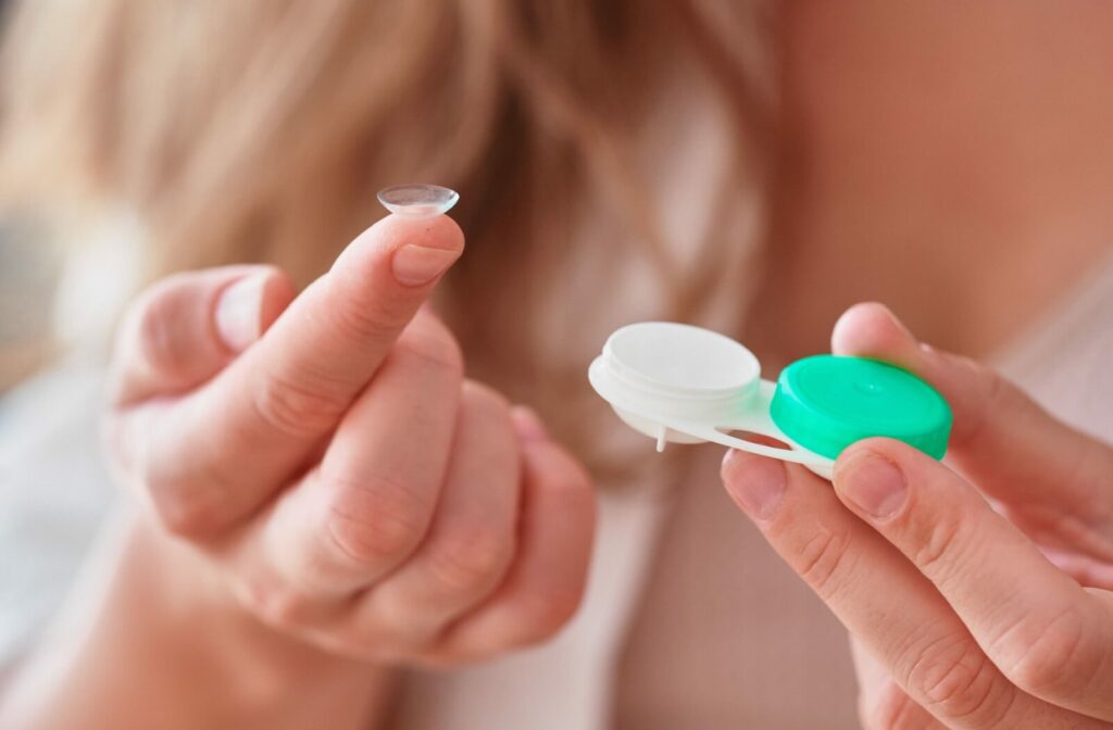 A pointer finger holds a soft contact lens while the other hand holds the contact lens storage container.