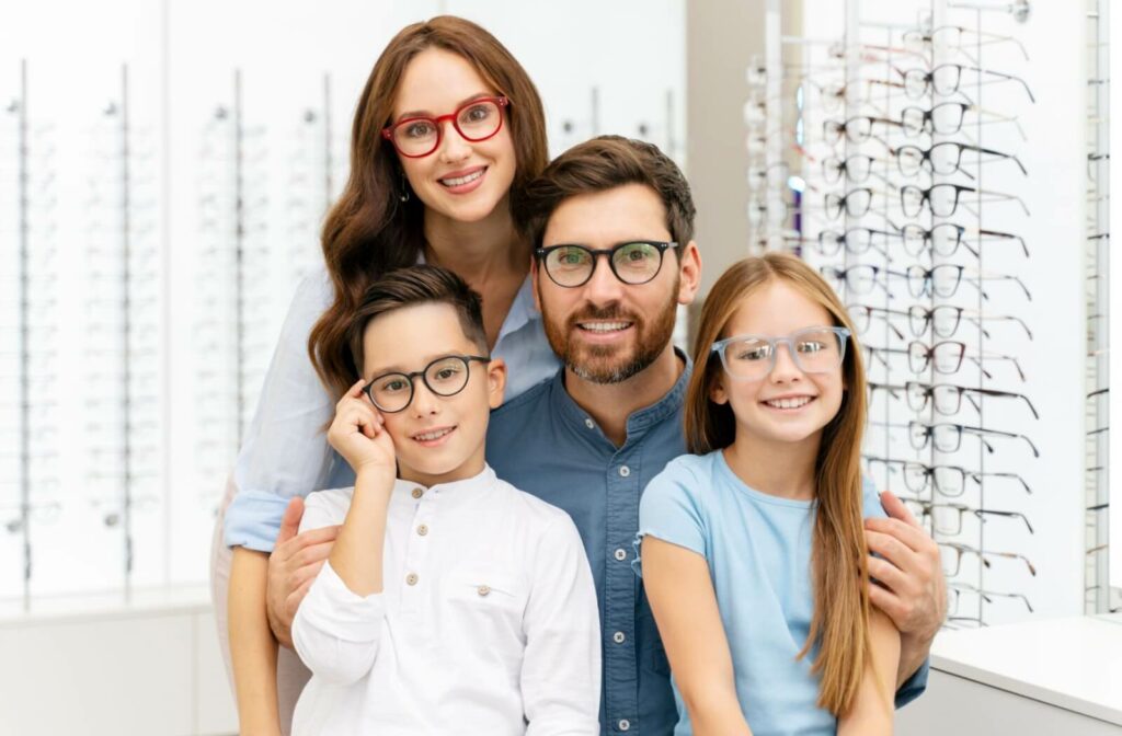 Smiling family of four wearing eyeglasses inside an optical store, standing in front of display shelves with frames