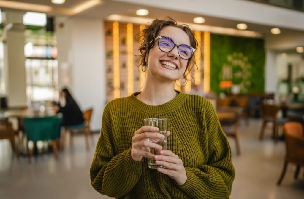 Person wearing glasses and smiling while holding a glass of water in a bright, modern indoor space.