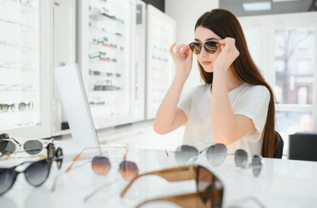 A person trying on sunglasses in an optical store, looking into a mirror with multiple pairs of sunglasses displayed on the counter in front of them.