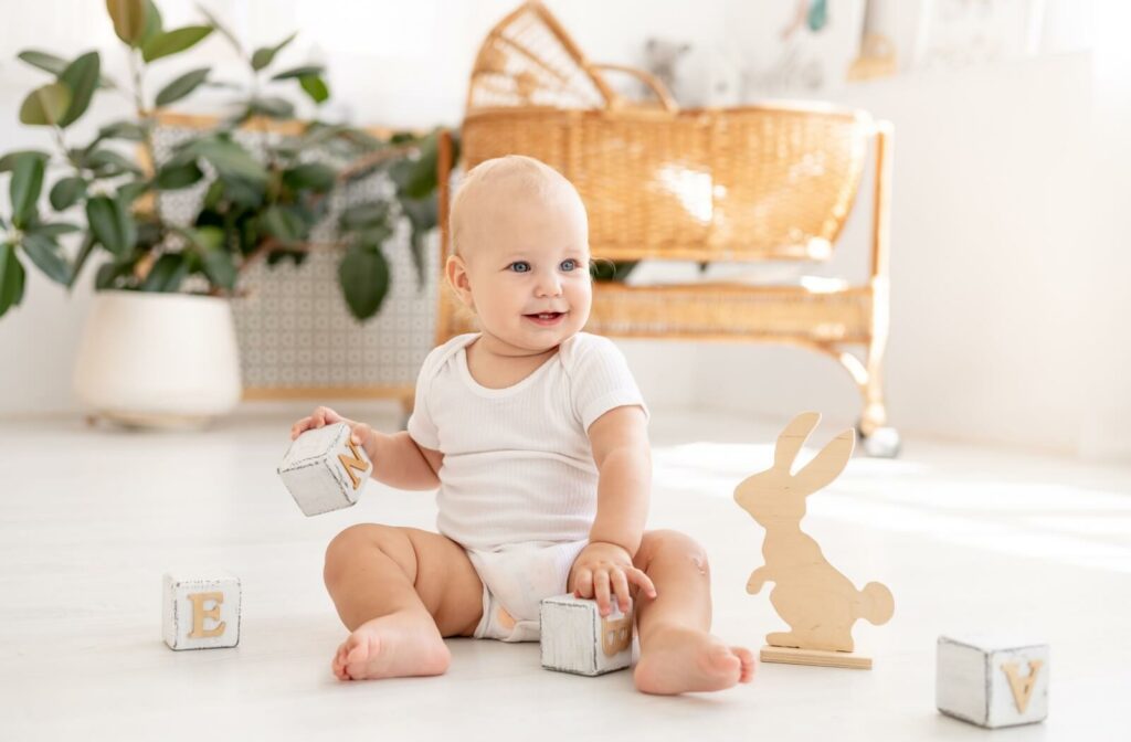 A smiling baby with blue eyes sitting on the floor playing with wooden blocks and a rabbit cutout.