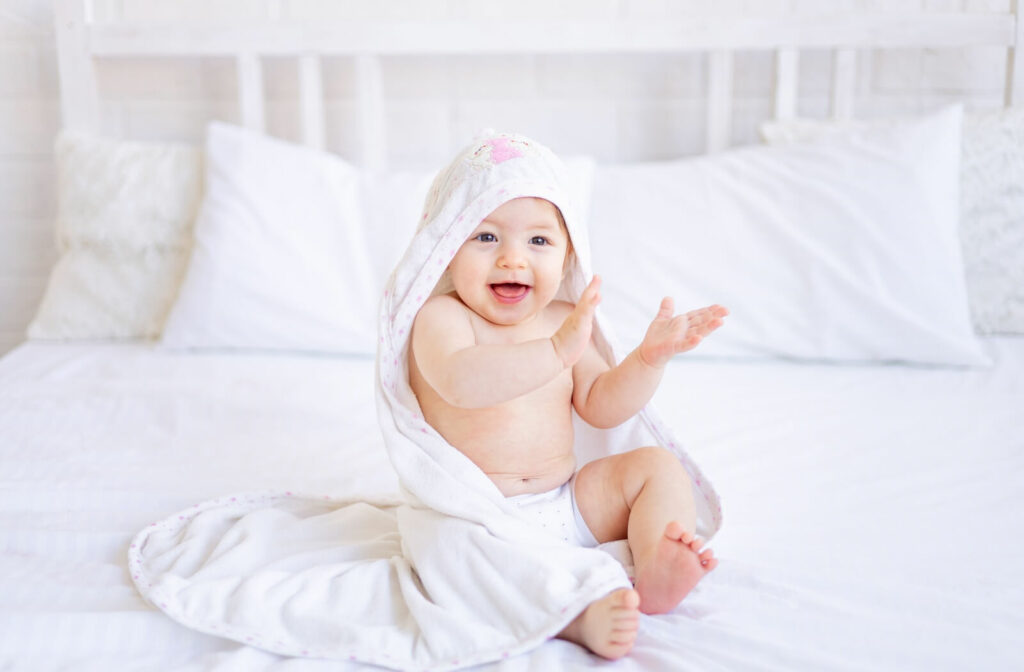 A happy infant sitting on a bed and clapping while wrapped in a hooded towel.