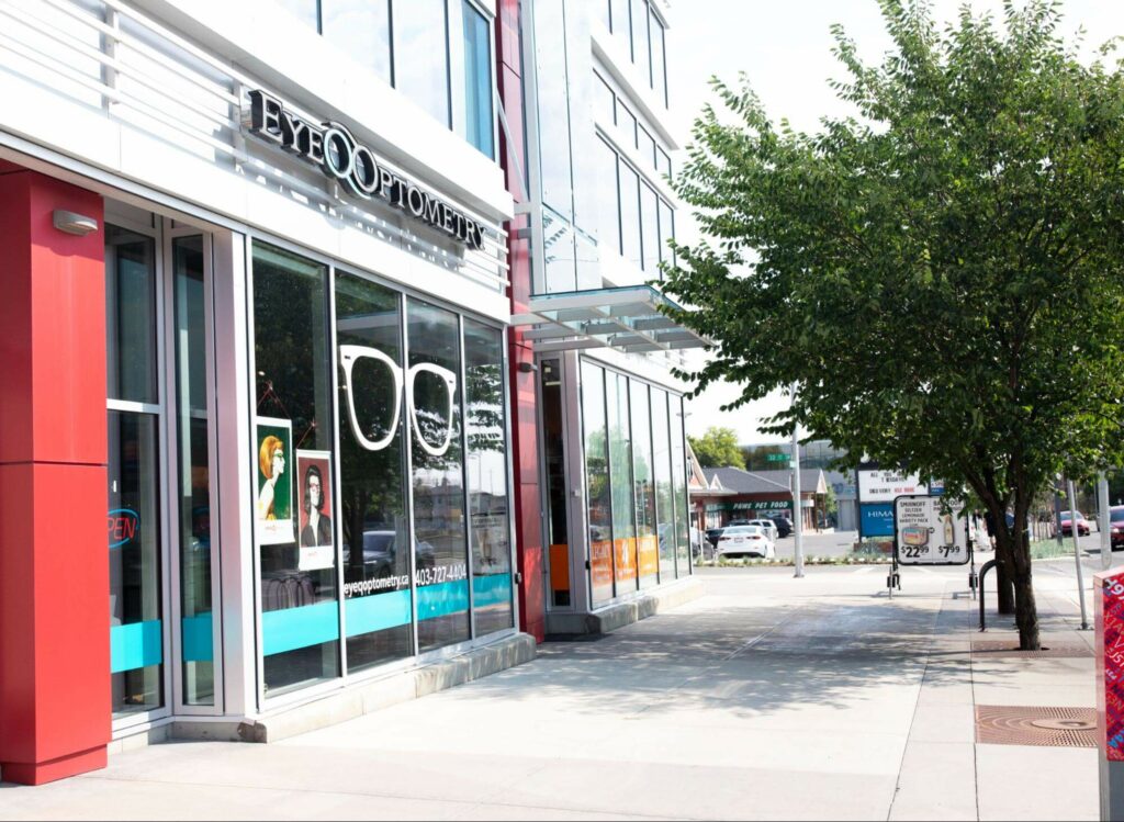 The modern exterior storefront of Eye Q Optometry in Calgary, featuring large glass windows with a white eyeglasses decal, red pillars, and a clean concrete walkway.