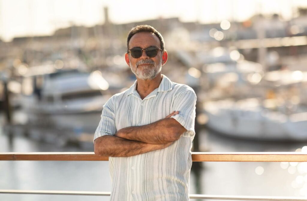 A senior man with a beard wearing sunglasses and a light-colored shirt, standing with arms crossed at a sunlit harbor to protect his eyes from UV glare.