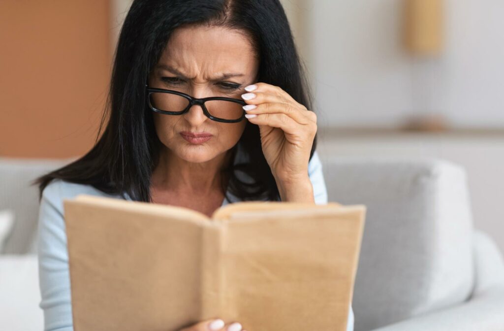 A middle-aged woman with dark hair frowning and peering over her reading glasses to clear up blurry text in a book, demonstrating common signs of presbyopia.