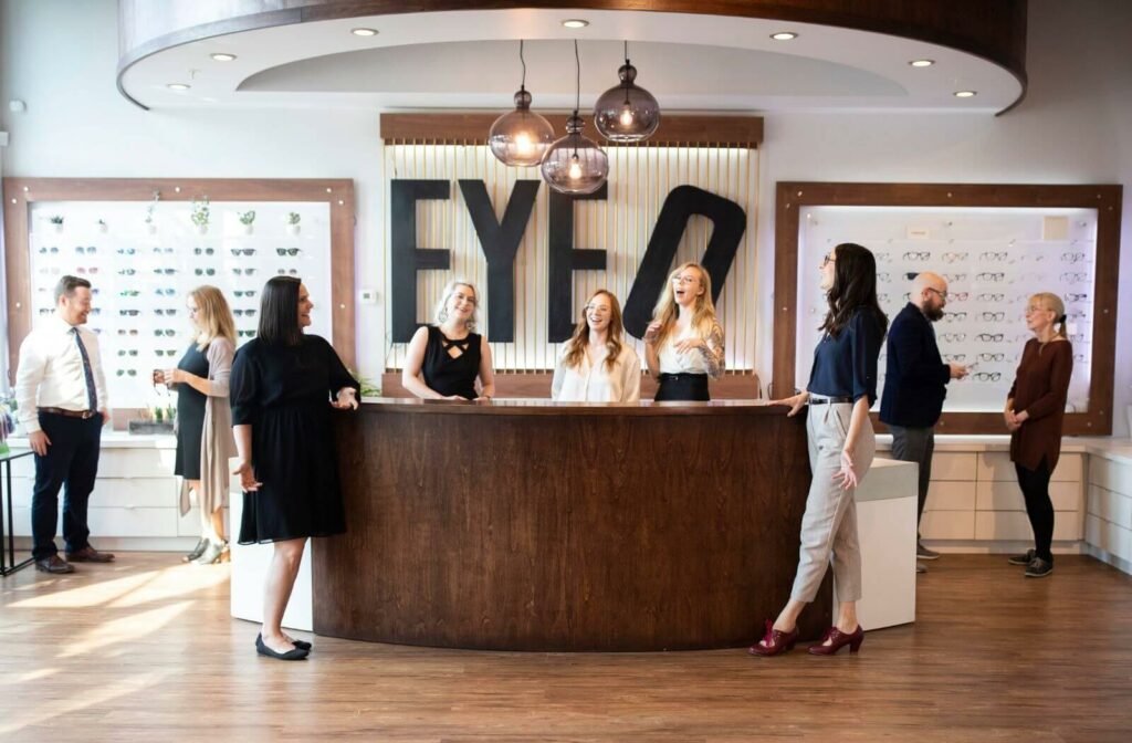 Optical store interior with staff and customers gathered around a reception desk, surrounded by eyewear displays.
